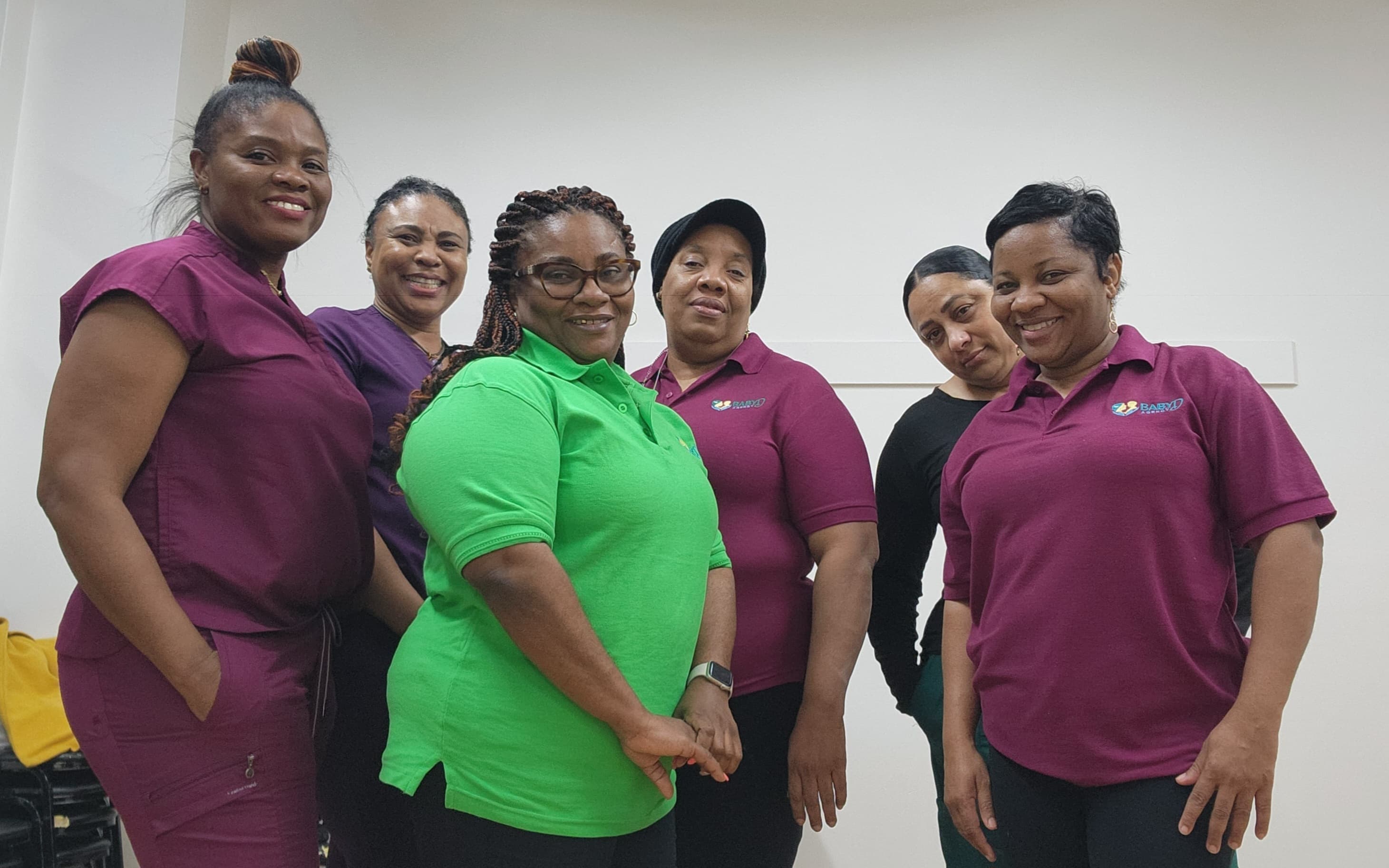 Six diverse women smiling together, wearing maroon scrubs and a bright green polo shirt.