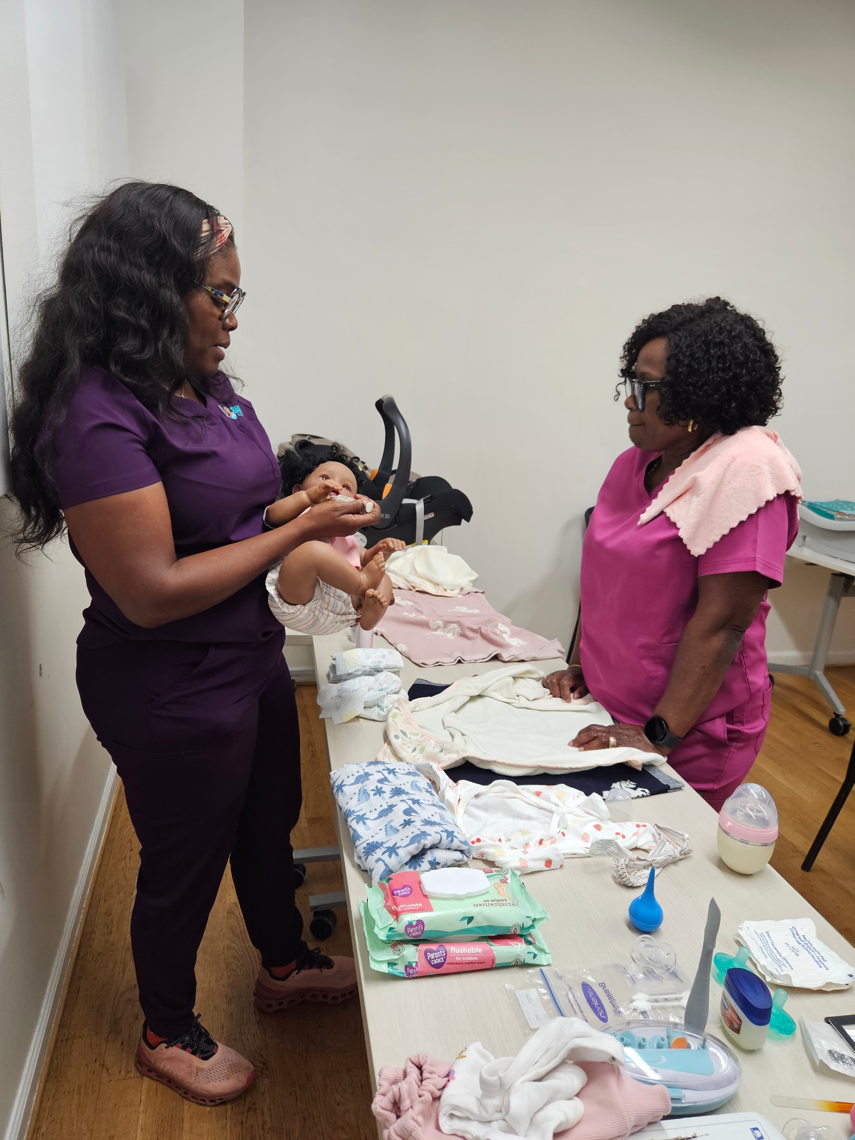 Two women in scrubs demonstrate newborn care with a baby doll and assorted baby supplies.