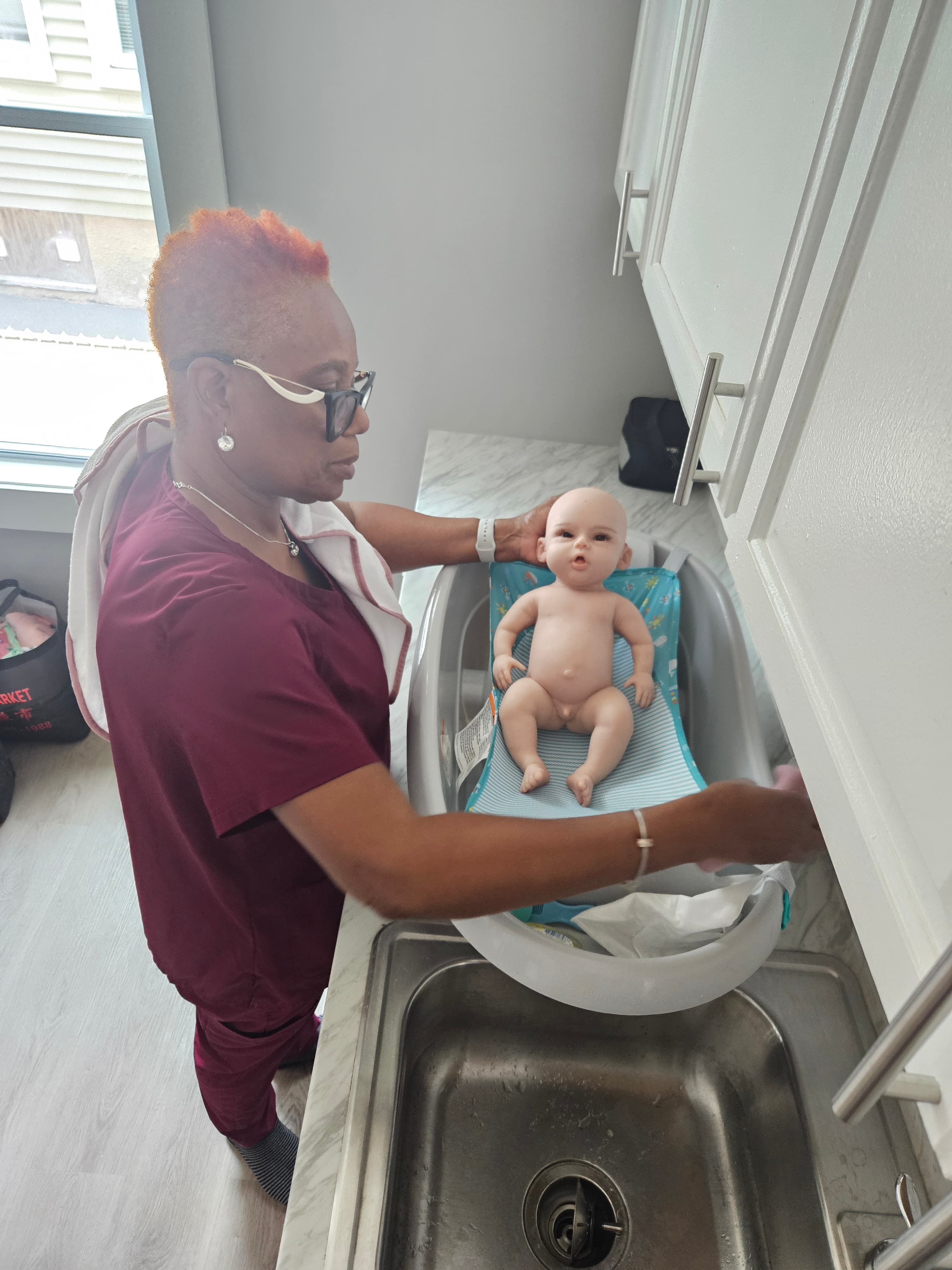 Woman in maroon scrubs leans over a baby doll in a small plastic bathtub.