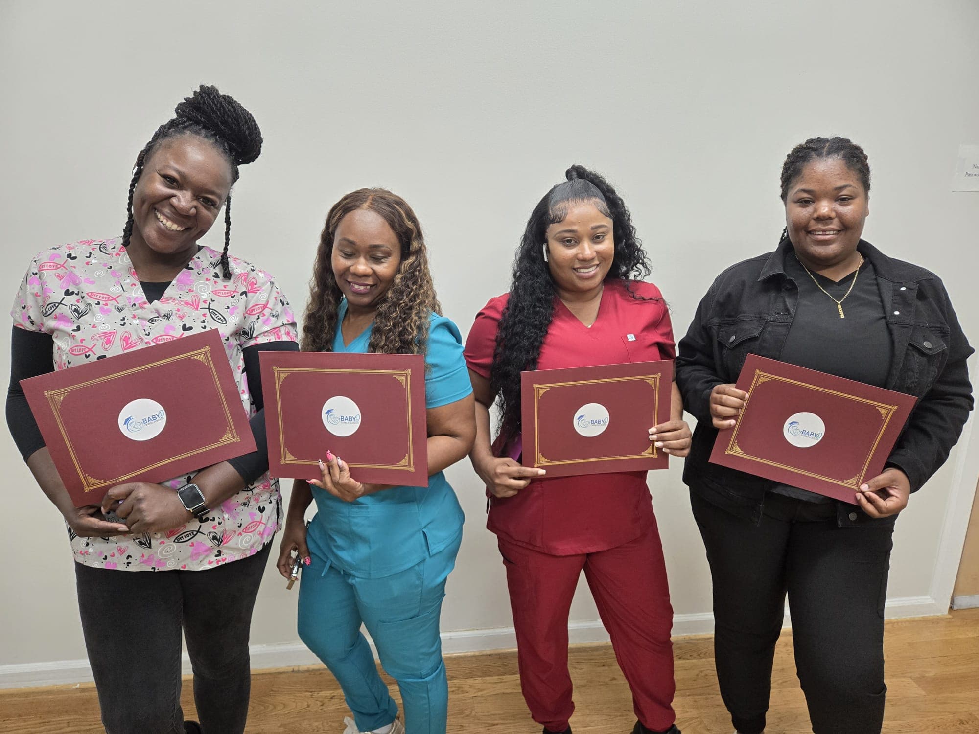 Four smiling women holding maroon certificates, three wearing medical scrubs, against a neutral wall.
