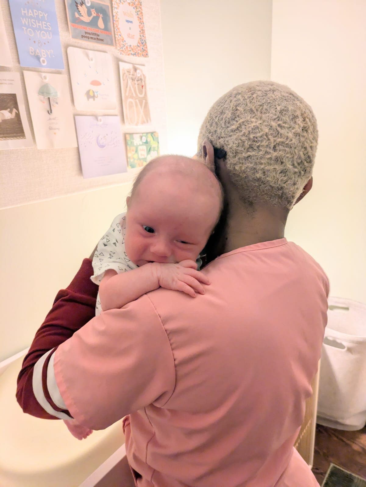 Person in pink scrubs holds a baby over their shoulder in front of greeting cards.
